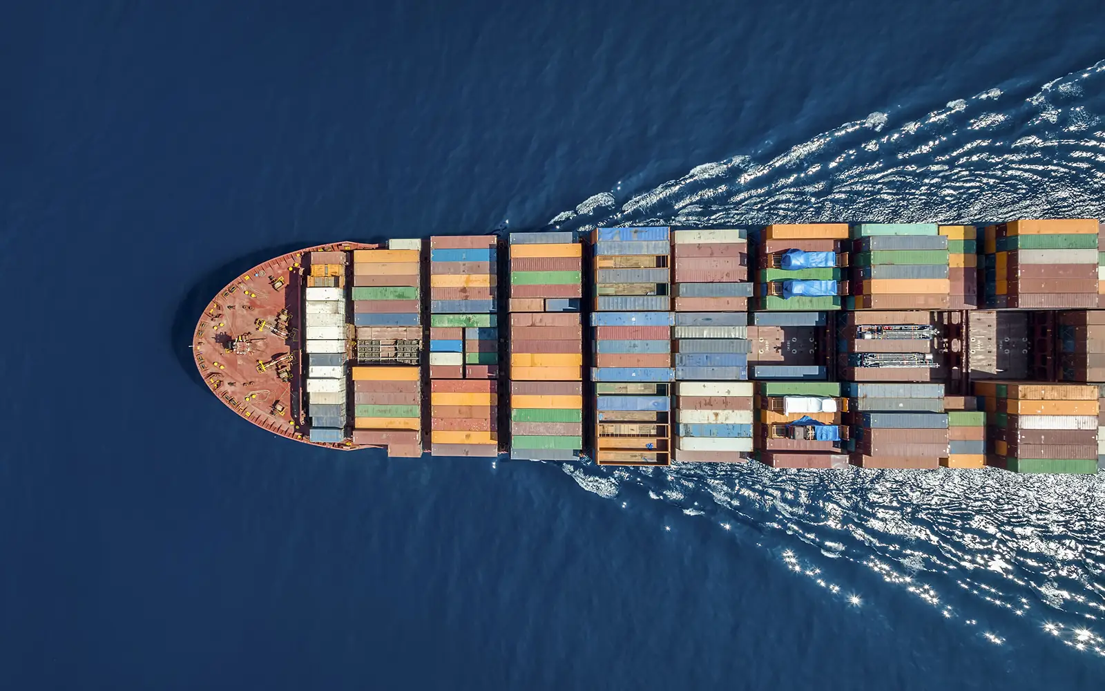 Aerial view of a large cargo ship navigating through deep blue ocean waters, loaded with colorful shipping containers, symbolizing global trade and supply chain logistics.